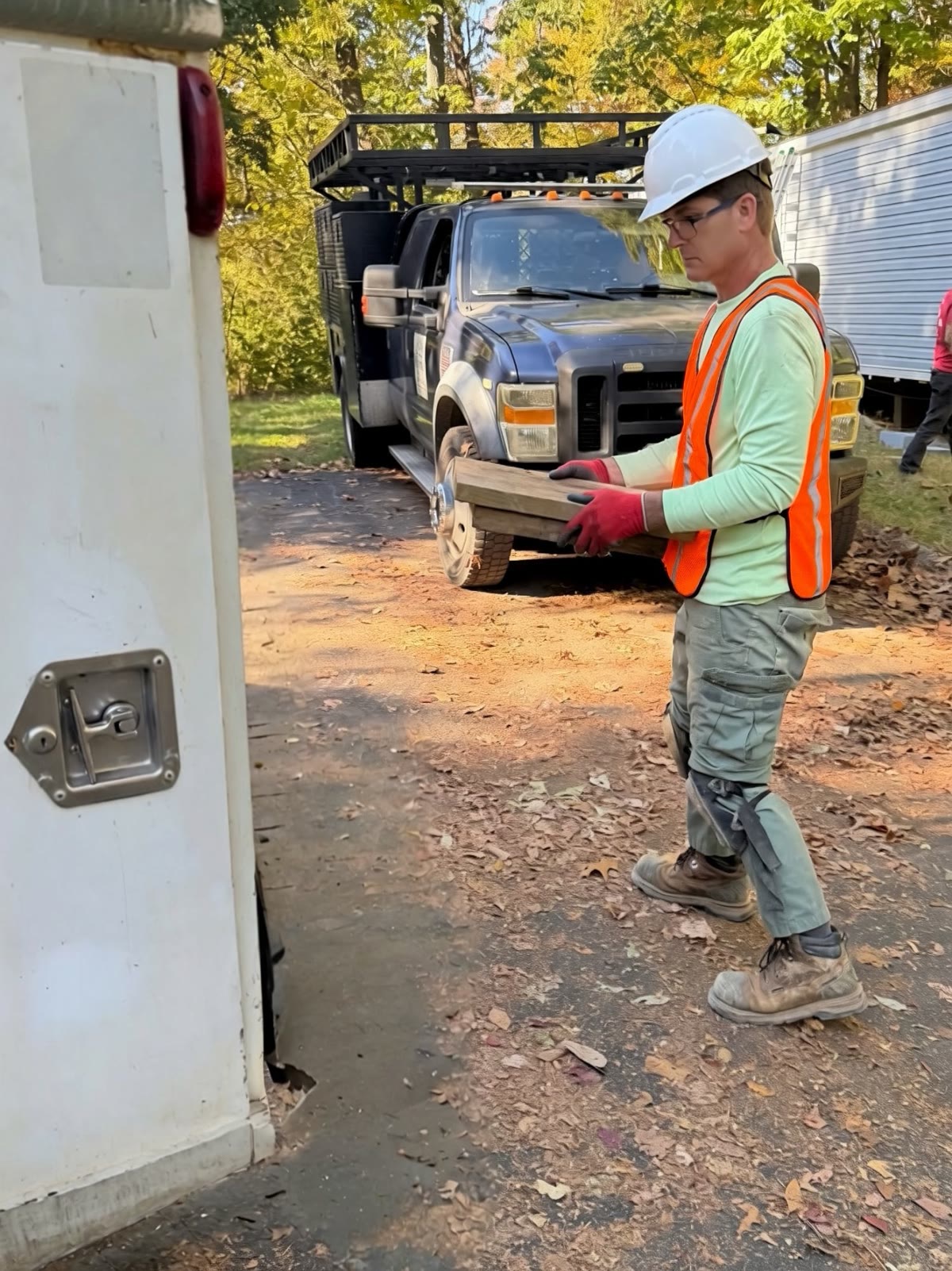 Crew member in a hard hat, hi-vis vest, and work gloves carrying wood blocking past a service truck